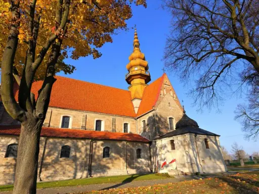 Kamienna świątynia z pomarańczowym dachem i żółtą wieżą, otoczona jesiennymi drzewami. To kościół św. Zbigniewa.