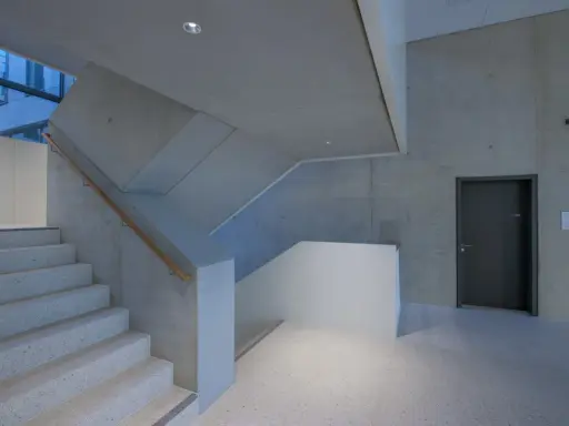 Moderne Treppe mit Sichtbeton-Wänden, die durch die Schalung eine interessante Textur erhalten. Ein dunkler Türrahmen und ein Holzhandschuh ergänzen das Bild.