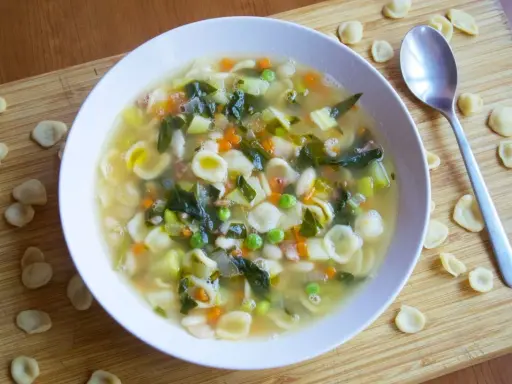 Un reconfortante plato de sopa minestrone, lleno de pasta orecchiette, verduras y frijoles, servido en un tazón blanco sobre una tabla de madera.