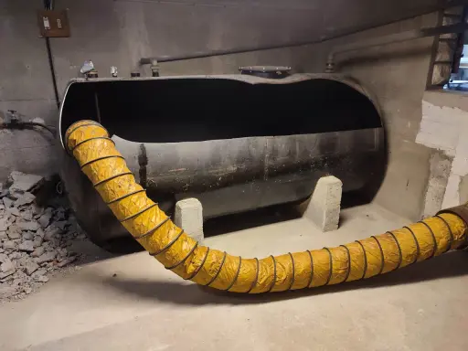 Gros réservoir de fioul noir, vide, avec un tuyau jaune flexible. Projet ma prime rénov cuve fioul en cours.