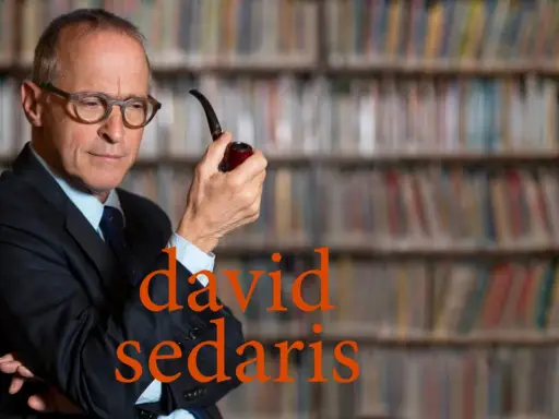 David Sedaris, con pipa in mano, seduto tra scaffali pieni di libri, riflette pensieroso.