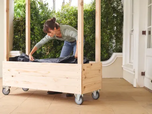 Une femme installe une bâche noire dans une jardinière en bois à roulettes, étape clé pour comment fabriquer toit pour tomates.