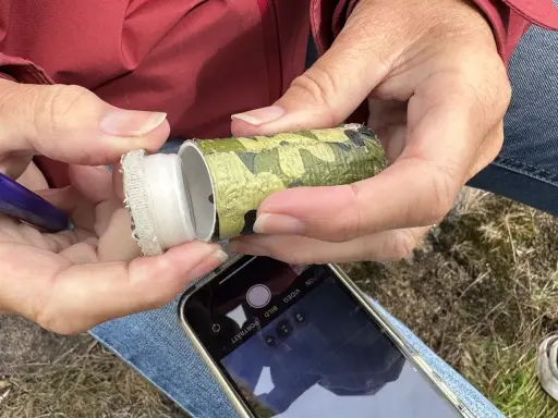 Händer öppnar en kamouflerad behållare, redo för geocaching i Sverige. En mobiltelefon ligger redo att dokumentera fyndet.