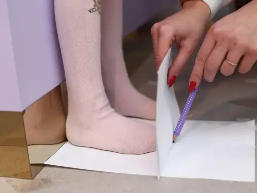 Dziecko mierzy stopę, by sprawdzić, ile luzu w butach będzie potrzebne. Rysunek ołówkiem na papierze.
