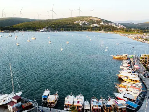 Port w Alacati, Turcja, pełen łodzi i żaglówek, z wiatrakami na wzgórzach w tle.