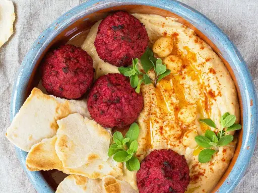 Rote Bete Falafel auf cremigem Hummus, garniert mit Kichererbsen und frischen Kräutern, serviert mit Pitabrot.