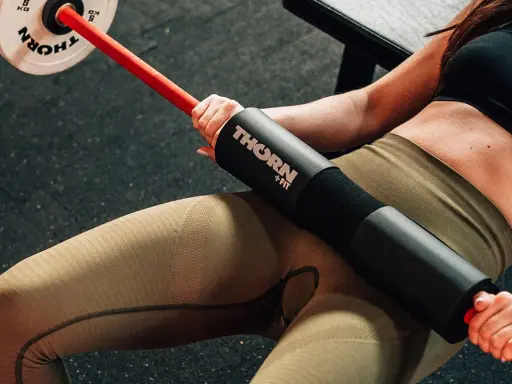 Kobieta wykonuje hip thrust w domu z gryfem i obciążeniem.