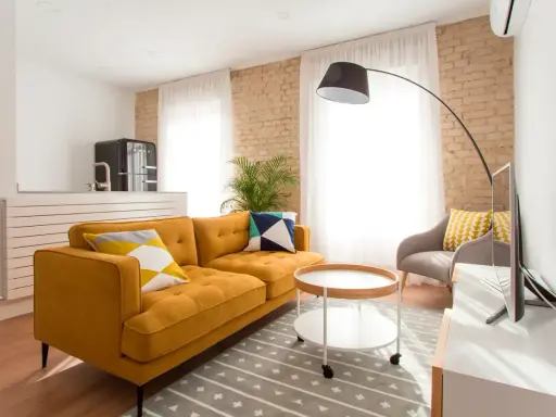 Sofá mostaza con cojines geométricos, mesa auxiliar redonda y alfombra gris. Los colores que combinan con el mostaza resaltan en esta sala.