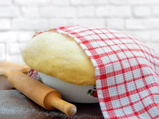 Wyrośnięte ciasto drożdżowe, przykryte ręcznikiem w kratkę, czeka na swój przepis babci. Obok wałek i mąka.