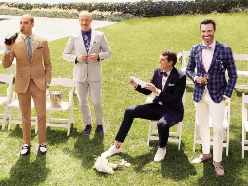 Four men in stylish garden formal wedding attire for male guests pose on a lawn. One drinks champagne, another adjusts his suit, one sits with a bouquet, and the last holds a drink with a dog nearby.