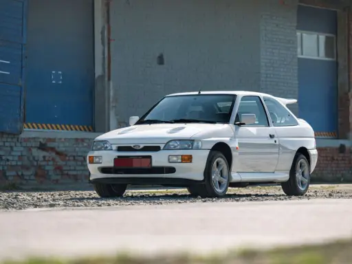 Biały Ford Escort Cosworth z charakterystycznym spojlerem, zaparkowany przed industrialnym budynkiem z niebieskimi drzwiami.
