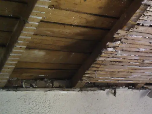 Vue intérieure d'une charpente en bois, montrant des lattes et des planches, avec des traces de plâtre. Un pare-pluie par l'intérieur semble avoir été appliqué.
