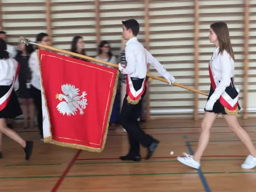 Uczniowie w galowych strojach tworzą poczet sztandarowy, niosąc biało-czerwony sztandar z godłem Polski.