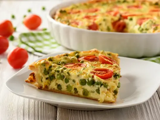 Ein Stück vegetarische Quiche mit Erbsen und Tomaten auf einem Teller. Das ganze Gericht ist im Hintergrund zu sehen.