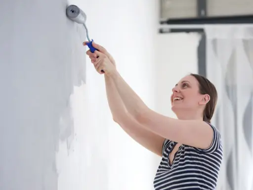 Une femme souriante apprend comment peindre un mur avec un rouleau. Elle applique une couche de peinture grise sur un mur blanc.