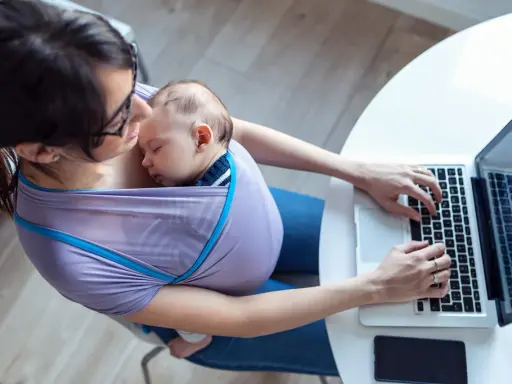 Kobieta w chuście nosi śpiące dziecko i pracuje na laptopie. Czy na macierzyńskim można pracować? Tak, to dowód.