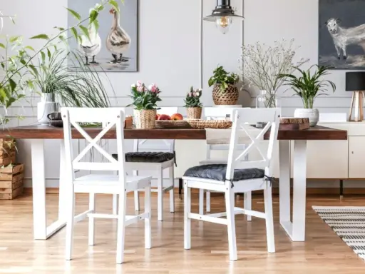 Comedor con mesa de madera oscura y sillas blancas, un ejemplo perfecto de cómo combinar muebles blancos y madera oscura. Plantas y cuadros decoran el espacio.