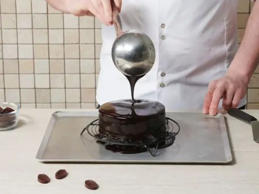Un chef verse un nappage chocolat brillant sur un gâteau. Des pépites de chocolat sont à côté.