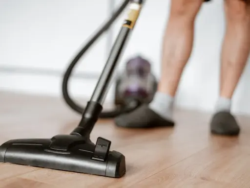 Odkurzacz na drewnianej podłodze, część nóg w kapciach i skarpetkach. To jeden z obowiązków domowych po angielsku – "vacuuming".