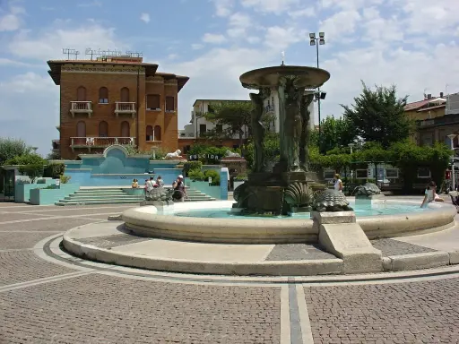 Fontana con statue e tartarughe in una piazza di Cattolica centro, con edifici storici e persone che si godono il sole.