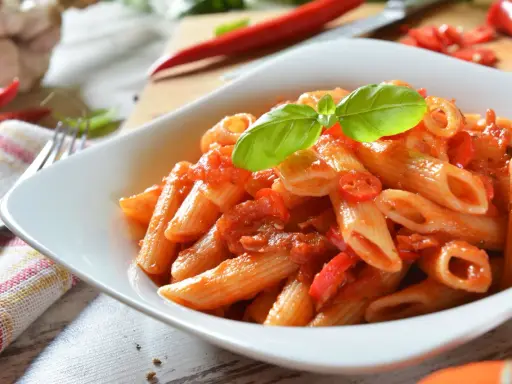 Eine Schüssel Penne Arrabiata, garniert mit Basilikum, mit scharfen Chilis und Knoblauch im Hintergrund.