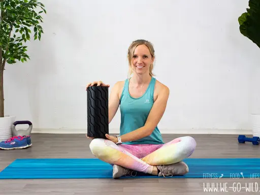 Frau zeigt, wie benutzt man eine Faszienrolle für das Training. Sie sitzt auf einer blauen Matte mit Gewichten und Pflanzen im Hintergrund.