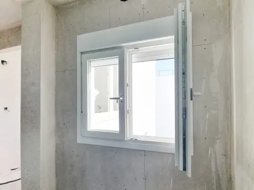 Instalar ventana PVC en obra gris. Ventana blanca de doble cristal con manilla plateada, lista para ser fijada.