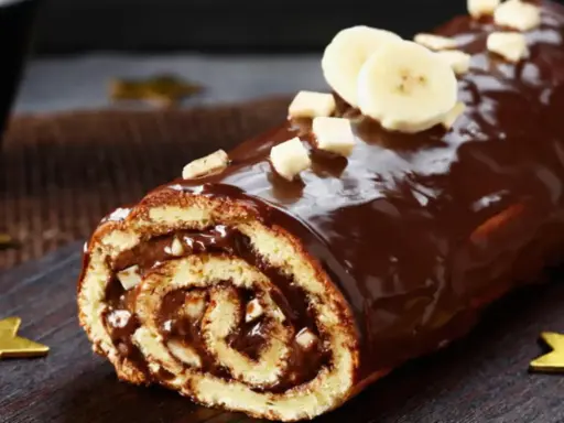 Bûche roulée au chocolat, garnie de crème et recouverte d'un glaçage chocolat beurre brillant, décorée de rondelles de banane et de noisettes concassées.
