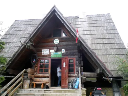 Drewniana bacówka PTTK pod Małą Rawką z otwartymi drzwiami, gdzie stoi mężczyzna. Obok stoją rowery i plecaki.