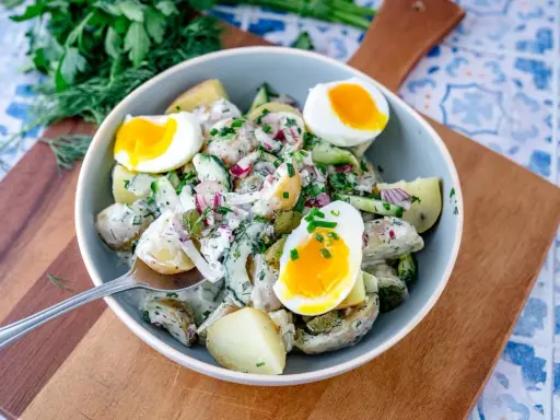 Schüssel mit cremigem Kartoffelsalat mit Gurken, Zwiebeln und gekochten Eiern, garniert mit frischen Kräutern.
