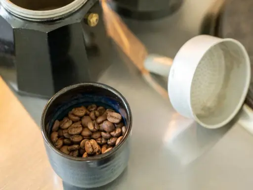 Kaffeebohnen in einer Schale neben einer Bialetti-Kaffeemaschine. Die Frage "bialetti wie viel kaffee" wird durch die Zubereitung beantwortet.