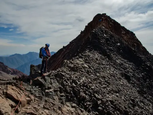 Alpinista na grani, zdobywający najwyższy szczyt Gruzji. W tle skaliste pasmo górskie i błękitne niebo.