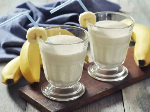 Dwa bananowe koktajle proteinowe w szklankach z rurkami i plasterkami banana. Idealne, gdy pić shake proteinowy po treningu.