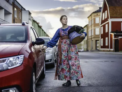 Lina Odell, klädd i folkdräkt, öppnar bildörren med en korg i famnen.