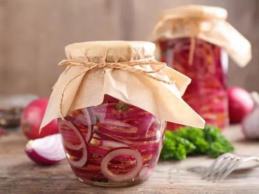 Zwei Gläser mit eingelegten Zwiebeln, verziert mit Papier und Schnur. Daneben liegen frische Zwiebeln und ein Gabel.