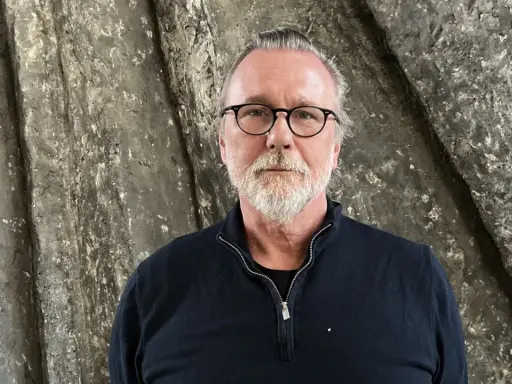 Thomas Olsson, en man med glasögon och grått skägg, står framför en grov, stenliknande vägg.