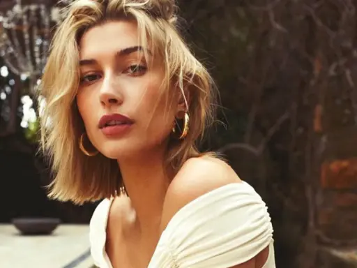 Mujer con un moderno corte bob rubio, aretes dorados y un vestido blanco con hombros descubiertos, posando al aire libre.