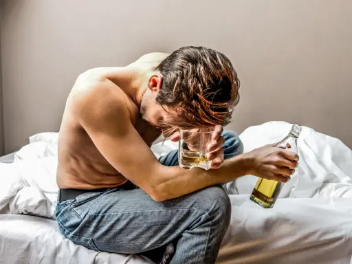 Mężczyzna siedzi na łóżku, trzymając szklankę z alkoholem i butelkę.