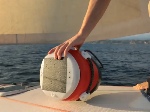 Main d'un marin tenant une bouée orange et blanche, un câble y est accroché. Le bateau navigue sur une mer bleue sous un ciel clair.