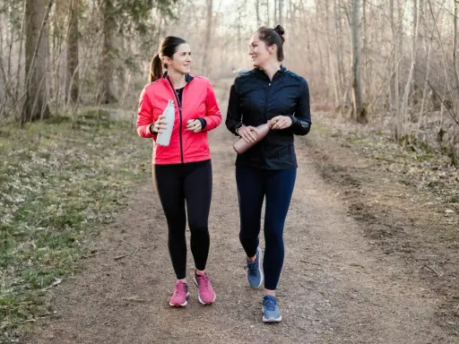 Two women in athletic wear walk on a forest path, holding water bottles.