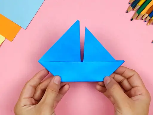 Hände falten ein blaues papierschiffchen. Bunte Stifte und Papier liegen bereit, um das papierschiffchen bauen zu beginnen.