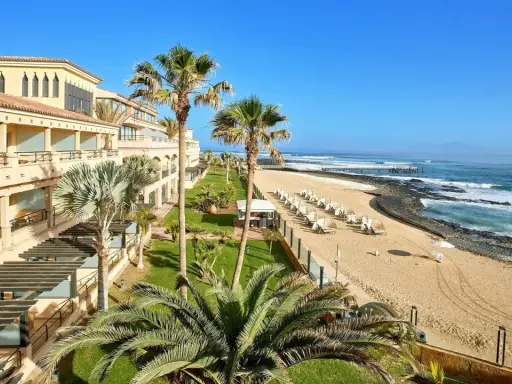 Fuerteventura kiedy wieje, palmy kołyszą się nad piaszczystą plażą i oceanem. Hotel z tarasami i widokiem na fale.