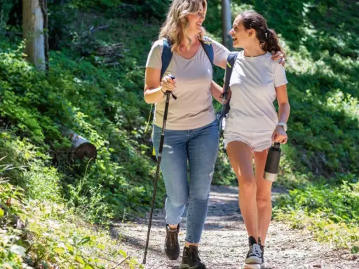 Två glada kvinnor njuter av en promenad i skogen, med en vandringsstav och en vattenflaska.