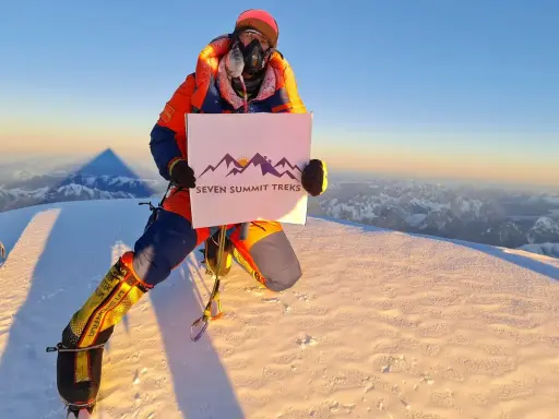 Na szczycie, gdzie jest K2, wspinacz w pomarańczowym kombinezonie trzyma tabliczkę "Seven Summit Treks".