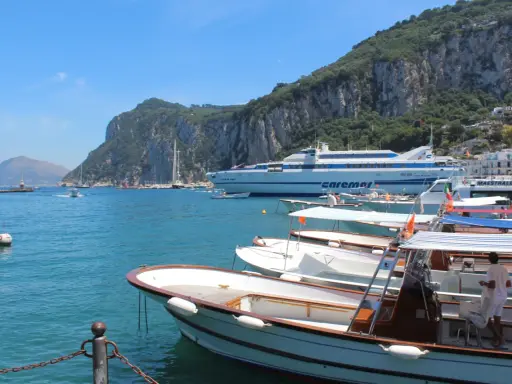 Barche in attesa al porto, con la maestosa isola di Capri sullo sfondo. Un traghetto Caremar attende passeggeri da Napoli.