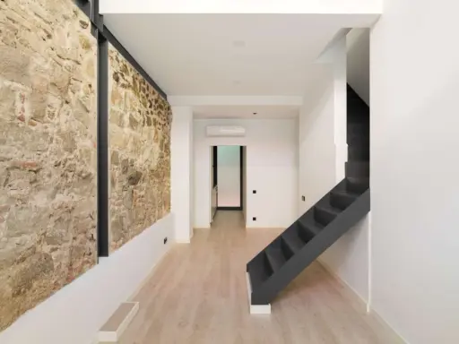Interior moderno con pared de piedra rústica, escaleras oscuras y suelo de madera clara. Un ejemplo de cómo hacer una casa sostenible combinando lo antiguo y lo nuevo.