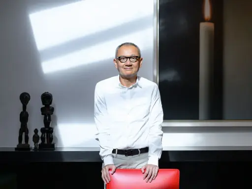 Pierre Chen, a man with glasses, stands in a modern room with striking shadows. He wears a white shirt and tan pants, leaning on a red chair.