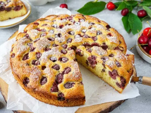 Saftiger Kirschkuchen mit frischen Kirschen, bestäubt mit Puderzucker. Ein Stück wird gerade herausgeschnitten.