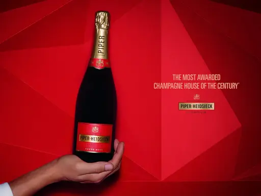 Eine Hand hält eine Flasche Piper-Heidsieck Champagner vor rotem, geometrischem Hintergrund.