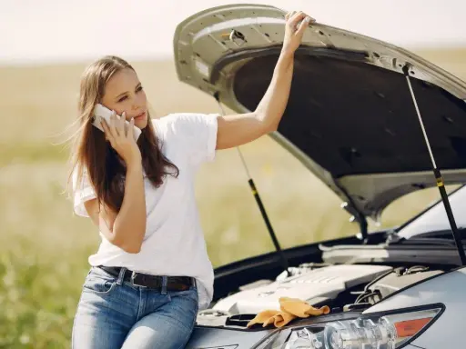 Mujer llama por teléfono con el capó abierto, preocupada por qué pasa si el coche se queda sin aceite.